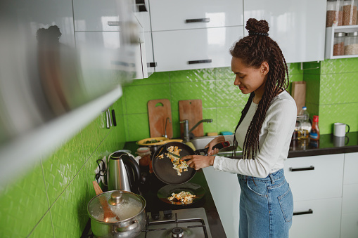 Happy mixed-race female cooking in kitchen in apartment serving food using frying pan, cook concept