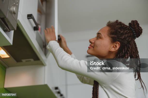 Joyful Female With Braids Opening Cupboard Indoor Woman Opens Kitchen Cabinet At Home Stock Photo - Download Image Now