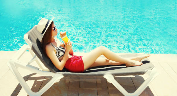 Summer vacation, happy relaxing young woman lying on deckchair with cup of juice on pool background stock photo