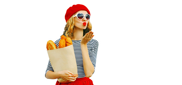 Portrait of beautiful woman blowing her red lips sending sweet air kiss holding grocery shopping paper bag with long white bread baguette wearing french red beret isolated on white background