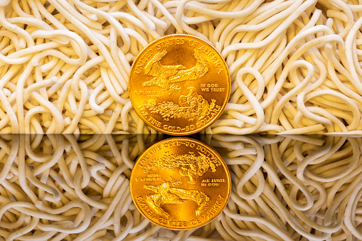 One ounce gold coin on reflective surface surrounded by egg noodles concept of food commodity inflation and hard currency barter in sanctioned global trade.