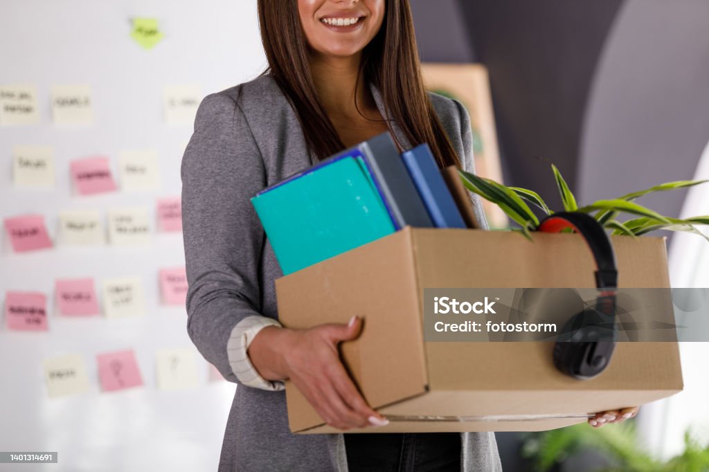 Joyful businesswoman smiling after getting hired at the office, holding a box with personal belongings Cut out shot of joyful young businesswoman smiling and holding a cardboard box with personal belongings after getting hired at the office. Quitting a Job Stock Photo