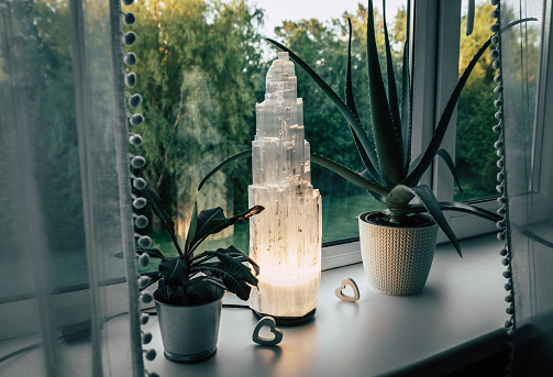 Rough big selenite crystal tower pole lamp illuminated in home on window sill, summer forest on background, spiritual home decor accent and ambient mood light concept.