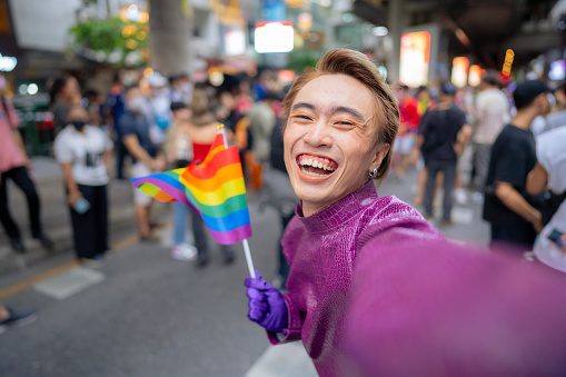 Confident Asian gay smiling and holding rainbow flag symbol of LGBTQIA social movement .
