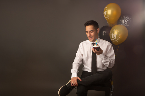 Smiling handsome mixed race man celebrating his birthday with cupcake and balloons in grey background studio.