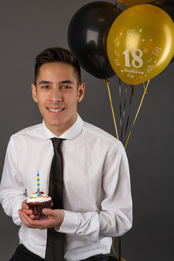 Handsome mixed race man celebrating his birthday with cupcake and balloons in grey background studio.