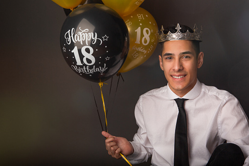 Cheerful andsome mixed race man celebrating his birthday with balloons sitting on grey background studio.