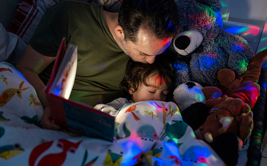 A directly above shot of a young boy wearing pyjamas lying in his bed with his father. His father is reading him a bedtime story out of a book.  There is a colourful night light projector that is projecting across the boys room, the father is kissing his son on the head as he's fallen asleep.