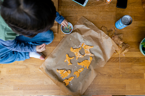 A directly above close-up shot of an unrecognisable young boy at a table at his home in the north east of England. He is preparing homemade cookie biscuits in the kitchen, they are on a baking tray and he's adding colourful sprinkles.
