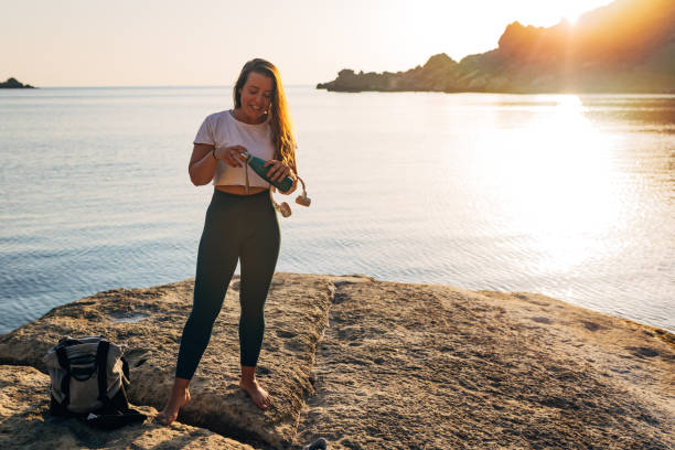 재사용 가능한 병에서 물을 마시는 바다 옆에 서있는 맨발의 여성의 정면 전망 - stone age audio 뉴스 사진 이미지