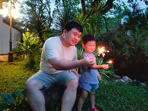 Asian toddler boy cuddling with his caring father who is holding a sparkler while they are celebrating New Year's eve.