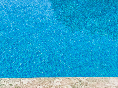 Geometric lines at the edge of a swimming pool with square mosaic pattern on floor. Marble frame. Crystal clear turquoise water background with idyllic liquid surface and wave pattern. Clean and transparent texture. Sunlight reflecting on ripples. Tropical summer vacation.