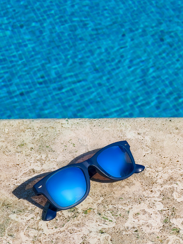A pair of blue mirror sunglasses at the pool side on sunny day. Sun reflecting on the glass. Summer vacation concept. Modern, single object exposed to sunlight. In background crystal clear turquoise swimming pool. Rippled water with wave pattern.