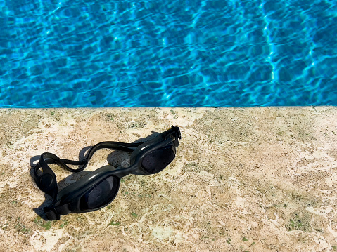 A pair of protective swimming eye wear on the side of a pool on sunny day. Summer vacation concept. Single object exposed to sunlight. In background crystal clear turquoise swimming pool water with wave pattern.
