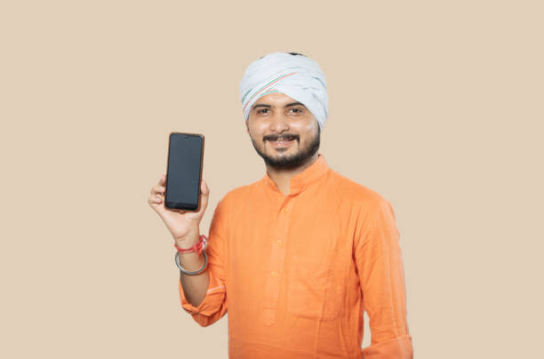 young indian rural mustache man wearing kurta and turban standing over isolated white background holding smartphone or mobile phone, village male with beard smiling showing cell phone, mock up. - kurta imagens e fotografias de stock
