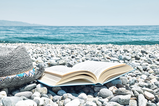 Open book and male straw hat on the pebble beach. Concept of reading and relaxing in summer vacation. Beach literature.