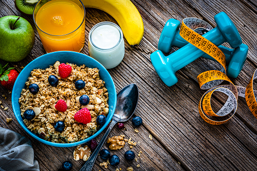 Healthy eating: overhead view of a blue bowl filled with granola shot on rustic wooden table. Dumbbells and a yellow tape measure are next to the bowl. A yogurt container, an orange juice glass, fruits and some nuts and berries are around the bowl. High resolution 42Mp studio digital capture taken with Sony A7rII and Sony FE 90mm f2.8 macro G OSS lens
