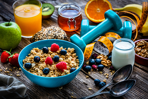 Healthy eating: blue bowl filled with granola shot on rustic wooden table. Dumbbells and a yellow tape measure are next to the bowl. A yogurt container, an orange juice glass, honey jar, fruits and some nuts and berries are around the bowl. High resolution 42Mp studio digital capture taken with Sony A7rII and Sony FE 90mm f2.8 macro G OSS lens