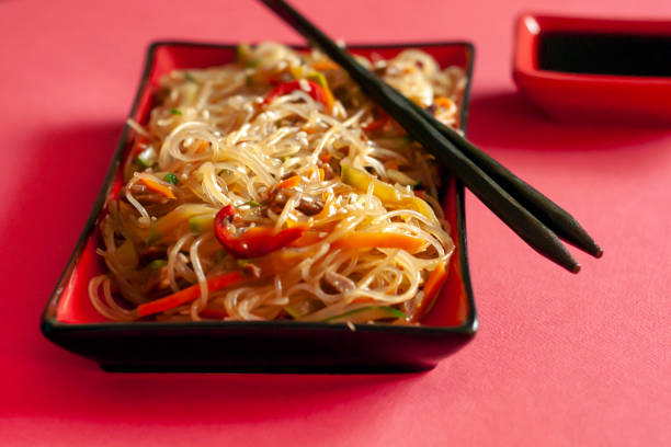 Traditional Korean starch noodles chapche with vegetables and beef in a red rectangular plate on a red background, chopsticks are on the plate, there is a saucepan with soy sauce next to it.