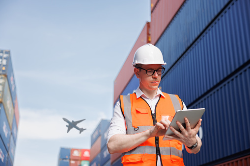 the logistic business owner is wearing a white safety helmet and using the digital tablet in a cargo freight ship for import export container yard with blue sky and airplane background.  the engineer and logistic planning manager.
