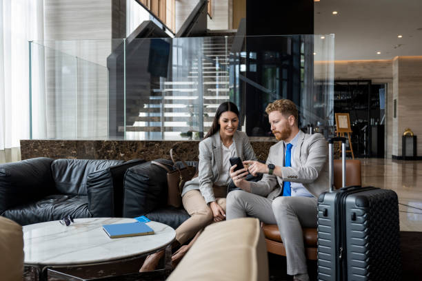 Hotel experience of two young colleagues Young businesswoman and her colleague seen in a hotel lobby sitting on the sofa and smiling while he is showing her something on his smart phone. business journey stock pictures, royalty-free photos & images