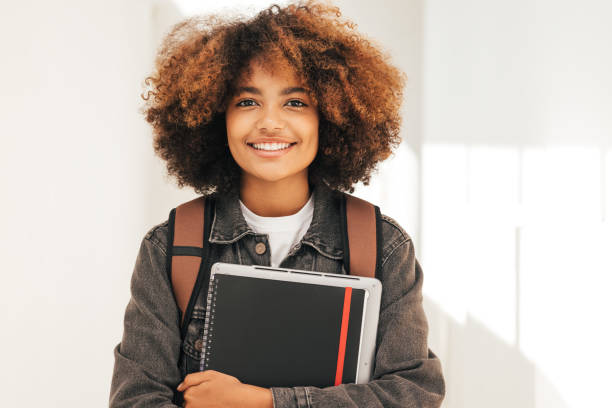 Close up portrait of a smiling student with textbook and laptop standing looking at camera stock photo