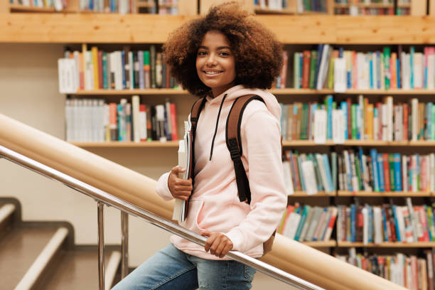 Beautiful girl with curly hair standing in library against bbokshelves and looking at camera stock photo