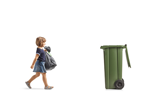 Full length profile shot of a little girl walking towards a dustbin and carrying a waste bag isolated on white background