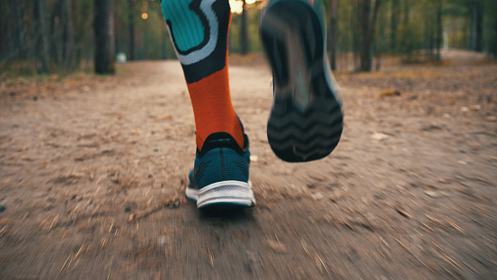Male feet in colorful long socks and blue sneakers run along forest path. Runner does training outdoors. Jogging through pine forest
