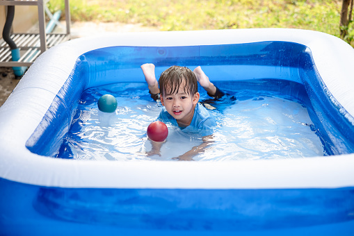 Portrait of asian child, kid or little boy to smile, bath, play water or funny activity in big blue portable inflatable swimming pool at outdoor, backyard or garden in summer evening. Include plastic color ball.