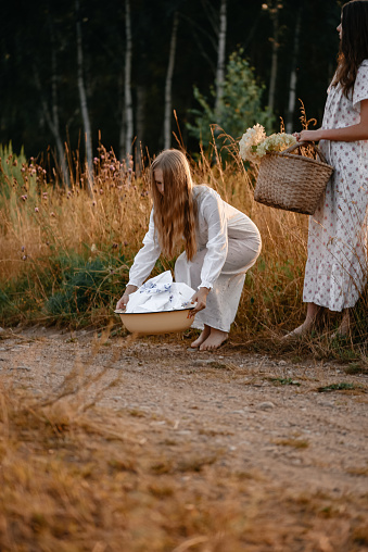 Two young girls in long white buds, posing at sunset near the field, a straw basket with flowers. The girl went out to wash things in the river. Basin with dirty linen. Evening walk in the village.