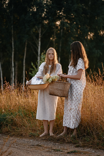 Two young girls in long white buds, posing at sunset near the field, a straw basket with flowers. The girl went out to wash things in the river. Basin with dirty linen. Evening walk in the village.