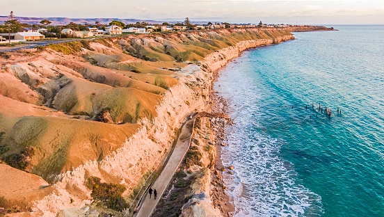 Aerial view Port Willunga sunset, clifftop houses with views of St Vincent Gulf, cliffs, rock face, ocean, unidentifiable couple in black walking on footpath, old jetty ruin and rolling hills in the distance.