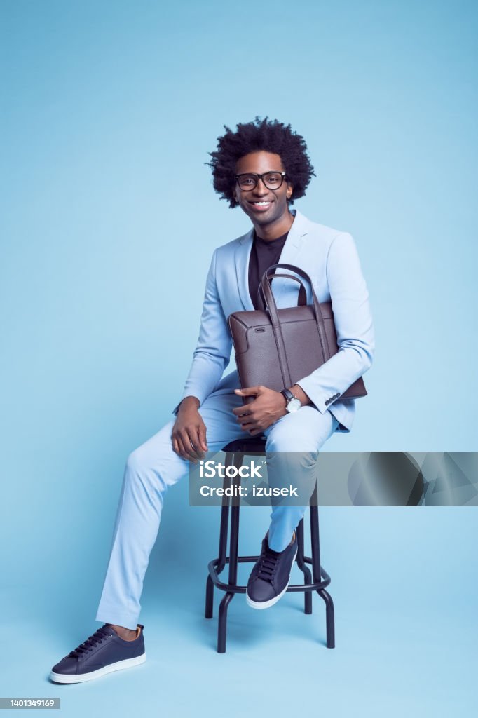 Man with briefcase sitting on stool Portrait of smiling businessman with briefcase sitting on stool against blue background Men Stock Photo