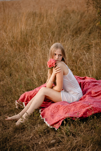 A young girl with long blond hair in a long white nightgown with flowers in her hands. Girl posing outdoors at sunset sitting on a blanket in a field. The girl collects wild flowers.