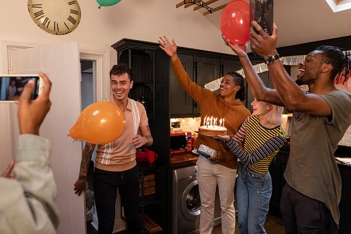 A small group of young adults surprise a friend by walking into the kitchen and greeting him with cake and balloons.