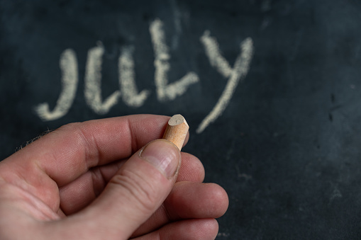 Adult male holding a piece of yellow chalk in his hand. Handwritten word JULY on black chalkboard. Selective focus.