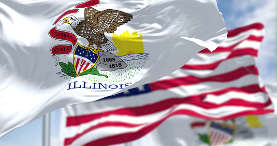 The Illinois state flag waving along with the national flag of the United States of America. In the background there is a clear sky. Illinois is a state in the Midwestern region of the United States