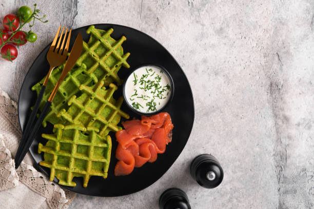 Green Belgian waffles. Spinach or wild garlic or pesto waffles with red salmon and cream sauce on grey concrete table background. Delicious breakfast, snack, brunch. Top view. Mock up stock photo