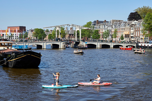 Two people paddleboarding with Skinny Bridge in the background on a summer day, Amstel River, Amsterdam, The Netherlands.