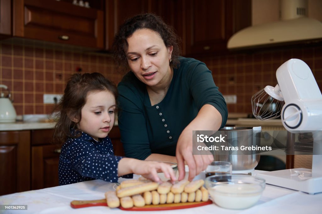 Multi-ethnic woman and Caucasian little girl- mom and daughter holding ladyfingers biscuits for prepare Italian dessert Tiramisu. Cooking at home, home food, home chef. Multi-ethnic woman and Caucasian little girl- mom and daughter holding ladyfingers biscuits for prepare Italian dessert Tiramisu. Cooking at home, home food, home chef and culinary concept. Baking Stock Photo