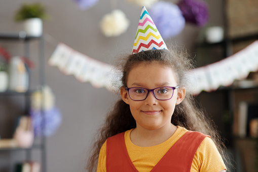 Portrait of little girl in eyeglasses and party hat smiling at camera at birthday party
