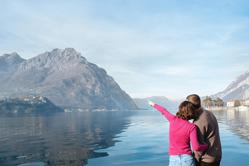 Soft focus, back view of couple enjoying a romantic date on the shores of Lake Lecco, Italy. The girl indicates direction with her hand. People in love on background of mountains, water. Copy space