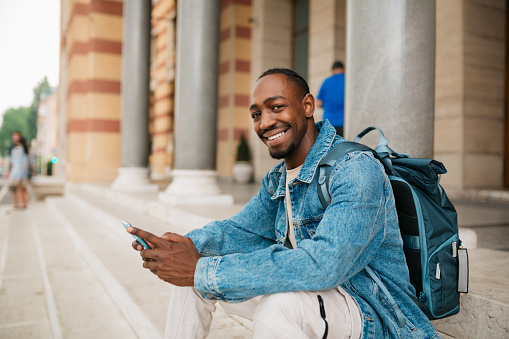 Portrait of a happy young adult male university student sitting on the steps in front of the university building and using a smart phone while looking at the camera