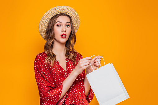 Portrait of a shocked surprised girl dressed in a red dress and straw hat with red lips holding shopping bags and looking at camera isolated over orange background