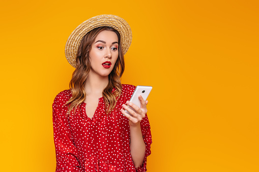 Surprised girl model in a red dress, red lipstick and a straw hat holds a mobile phone and reads an SMS message, makes online purchases, isolated on orange background, copy space. Summer girl with cell, sale concept
