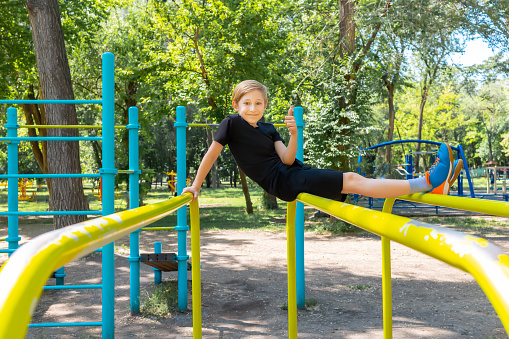 Cheerful boy in the park sits on gymnastic bars, laughs and shows a thumbs up
