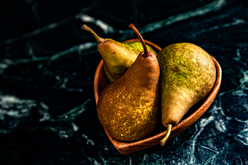 This is a moody lit photograph of three pears in a heart bowl on the marble green background
