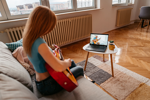 Young beautiful woman teaching to play acoustic guitar through a video call on laptop or watching tutorial.