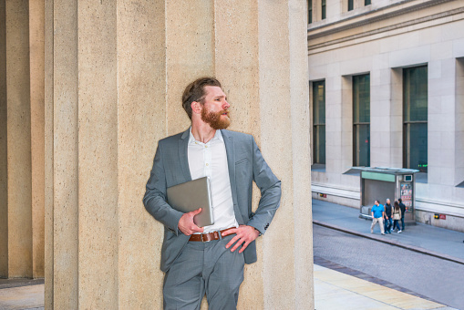 American Businessman with beard, mustache working in New York, wearing cadet blue suit, white undershirt, carrying laptop computer, standing against column outside office building, looking at street.